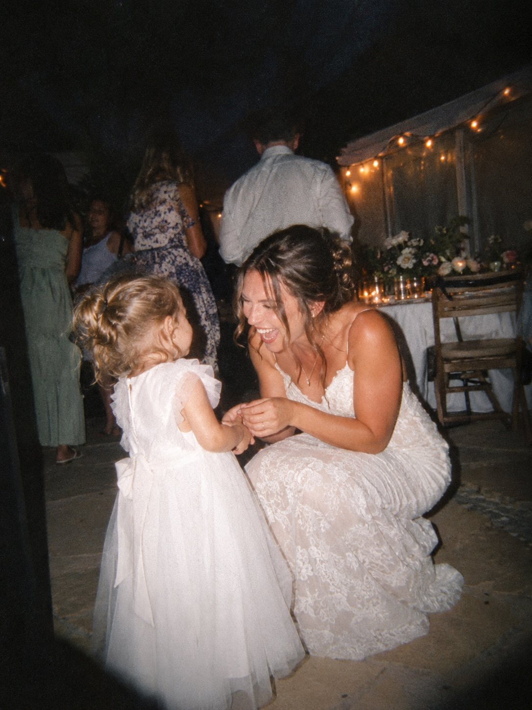 A bride kneels to hold hands and laugh with a little girl in a white dress, under string lights at an evening party.